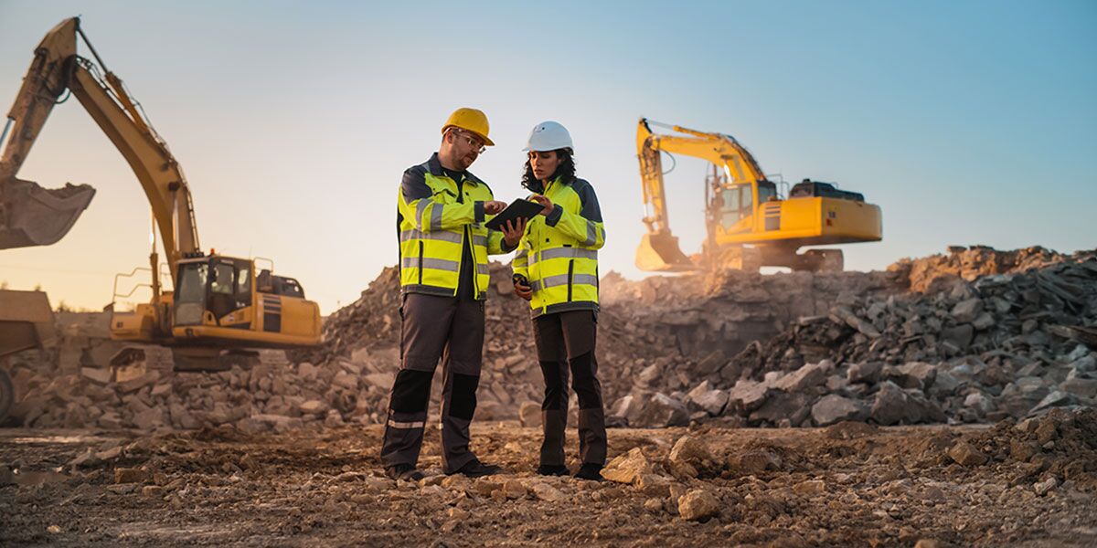 Two civil engineers stand at mine site in front of two excavators