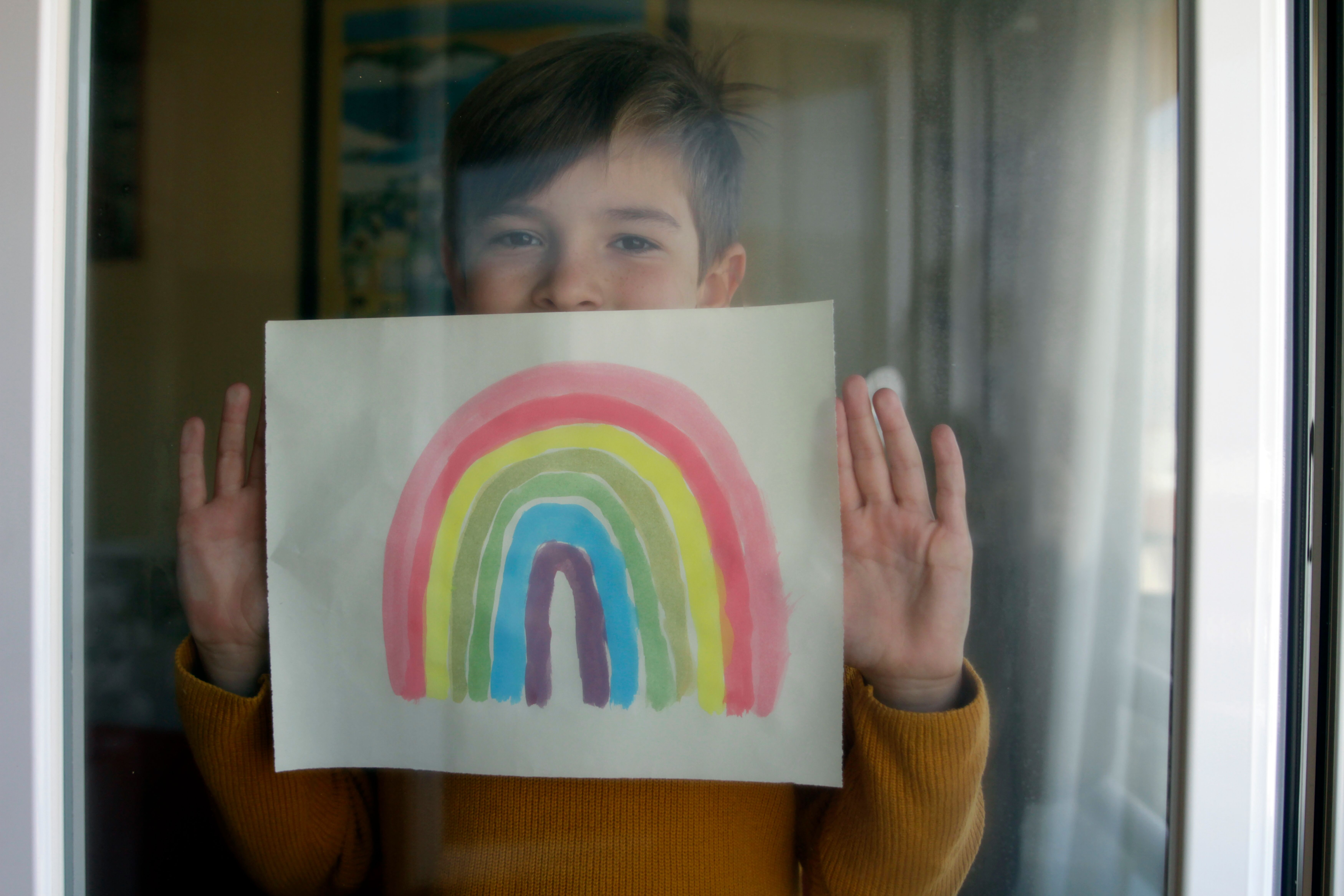 A young child holds up a painting of a rainbow to the window.