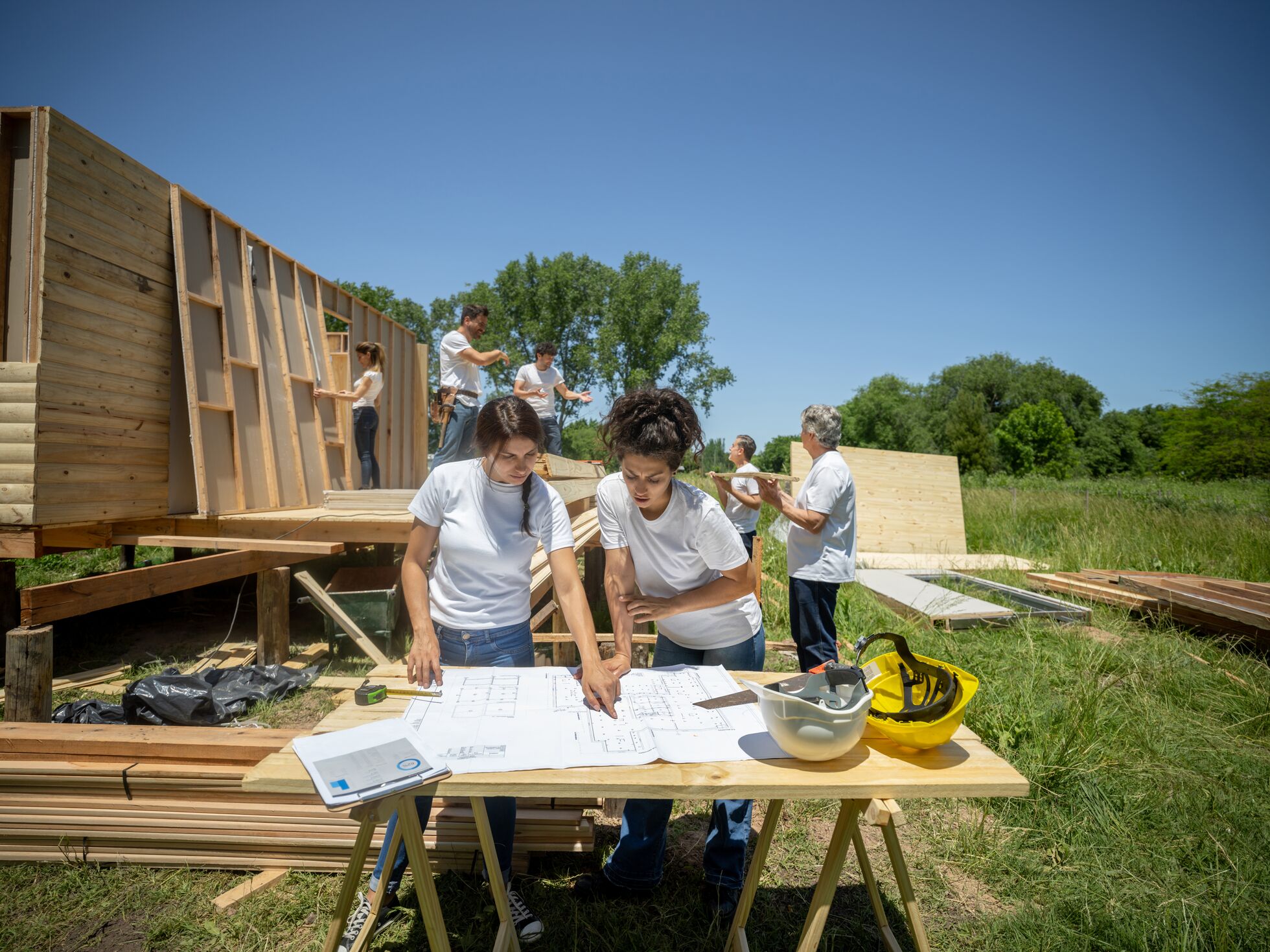 Two people observing plans at a temporary table while people in the background work on a house
