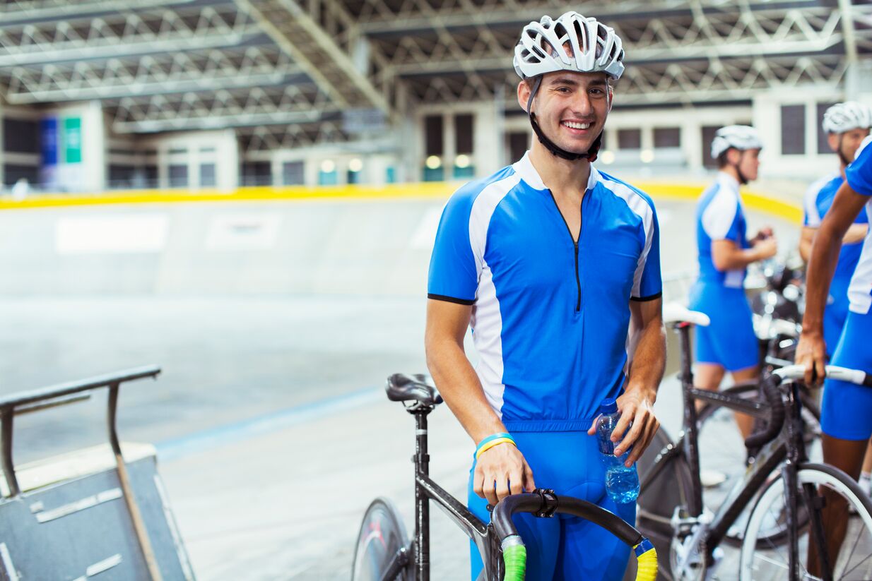 Cyclist wearing a blue jumpsuit and white helmet stands with his bike in a velodrome.